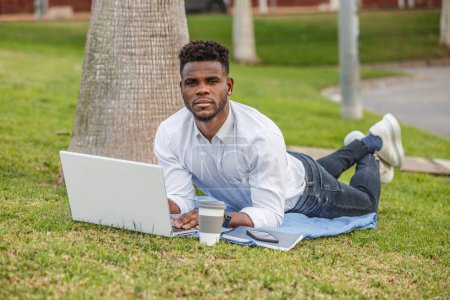 An African-American man is seen lying on the grass, using a laptop to work or study.