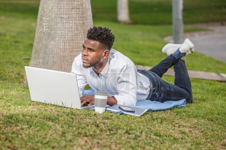 A man is seen peacefully lying on the grass while using his laptop for work or leisure.