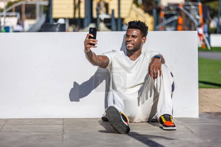 Foto de Un afroamericano de unos 30 años se sienta en el suelo en un parque público, capturando un selfie con su teléfono. - Imagen libre de derechos