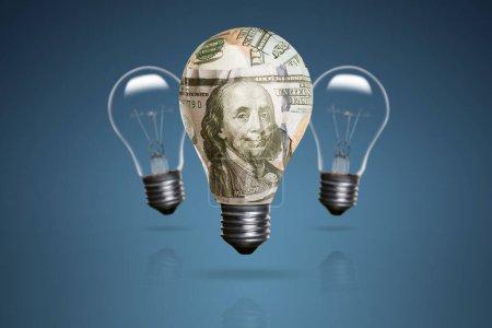 Foto de Una bombilla envuelta con unos billetes de 100 dólares y hay dos bombillas en el fondo y sobre un fondo azul. - Imagen libre de derechos
