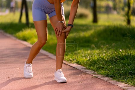 Téléchargez les photos : Gros plan du genou d'une femme sur une piste de course avec des collants bleus tenant son genou dans la douleur. - en image libre de droit
