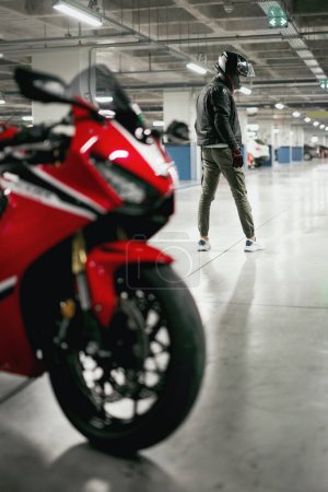 Foto de Vista trasera de un motociclista con un casco y una moto de carreras desenfocada en un aparcamiento. - Imagen libre de derechos
