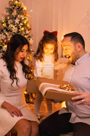 Glückliche Familie mit überraschenden Emotionen beim Öffnen einer Geschenkbox gibt es einen Weihnachtsbaum, Kamin und Kerzen auf dem Hintergrund.