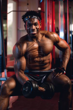 Photo for 20s Black and muscular man in a gym doing concentration curls with dumbbell and smiling expression - Royalty Free Image