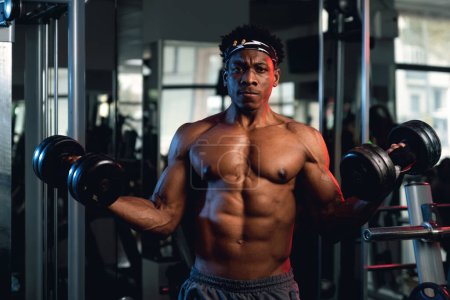 Photo for 20s Black and muscular man in a gym showing dumbbells and his muscles. - Royalty Free Image