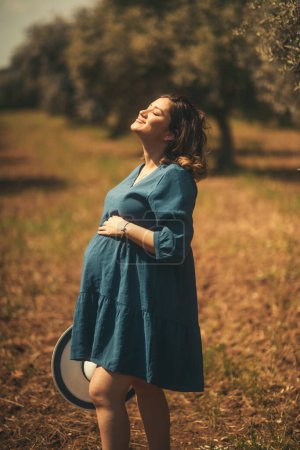 Téléchargez les photos : Une femme enceinte avec une robe bleue chapeau blanc dans un champ d'oliviers avec une faible profondeur de champ. - en image libre de droit
