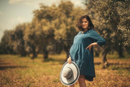 Téléchargez les photos : Une femme enceinte avec une robe bleue chapeau blanc dans un champ d'oliviers avec une faible profondeur de champ. - en image libre de droit