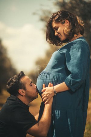 Téléchargez les photos : Un moment tendre comme un homme en t-shirt noir embrasse affectueusement le ventre de sa femme enceinte, tous deux debout dans un champ d'oliviers - en image libre de droit