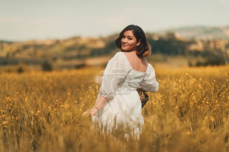 Téléchargez les photos : Une femme enceinte en robe blanche porte un panier de fleurs sèches dans un champ de blé doré, incarnant l'esprit de la fête des mères - en image libre de droit