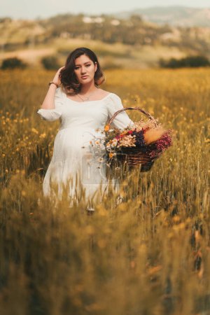 Foto de Una mujer embarazada con un vestido blanco lleva una cesta de flores secas en un campo de trigo dorado, encarnando el espíritu del Día de la Madre. - Imagen libre de derechos