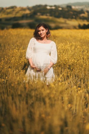 Téléchargez les photos : Une femme enceinte en robe blanche se tient élégamment dans un champ de blé doré, rayonnant de sérénité maternelle. - en image libre de droit