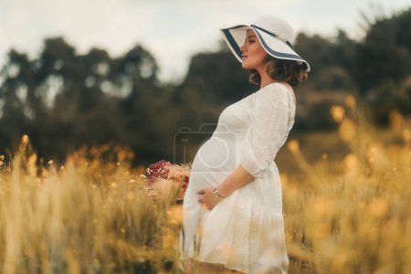 Foto de Encarnando el espíritu del Día de la Madre, una mujer embarazada con un vestido blanco y sombrero lleva una cesta de flores secas en un campo de trigo dorado. - Imagen libre de derechos
