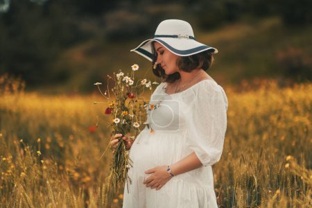Foto de Celebrando el Día de la Madre, una mujer embarazada con un vestido blanco y sombrero sostiene un montón de margaritas recién recogidas en un campo de trigo dorado - Imagen libre de derechos