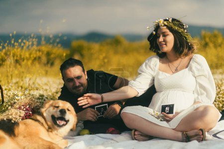 Foto de En una manta blanca de picnic, una mujer embarazada y su esposo comparten un momento tierno con la imagen de ultrasonido de su bebé, mientras que su amigable Golden Retriever busca afecto cerca - Imagen libre de derechos