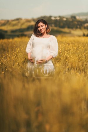 Téléchargez les photos : Une femme enceinte en robe blanche et chapeau se tient élégamment dans un champ de blé doré, rayonnant de sérénité maternelle. - en image libre de droit
