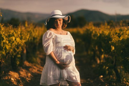 Foto de Retrato de una mujer embarazada vestida de blanco y sombrero, sosteniendo suavemente su vientre, en el marco de un viñedo, radiante maternidad y expectativa - Imagen libre de derechos