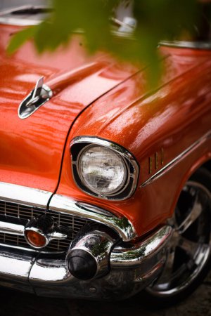 Foto de Izmir, Turquía - 3 de junio de 2023: Primer plano del faro empapado en lluvia y rueda parcialmente girada de un Chevrolet Impala naranja 1957, con hojas verdes en primer plano en el IZKOD Classic Car Meet en Buca Pond, Izmir. - Imagen libre de derechos