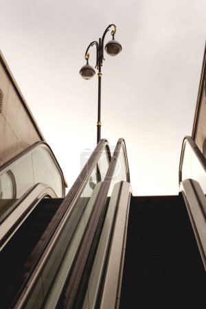 Téléchargez les photos : Cette photo capture un escalier roulant solitaire menant à un lampadaire, illustrant la solitude d'un paysage urbain. Sans personne en vue et avec un ciel nuageux au-dessus, la scène évoque un sentiment d'anticipation tranquille - en image libre de droit