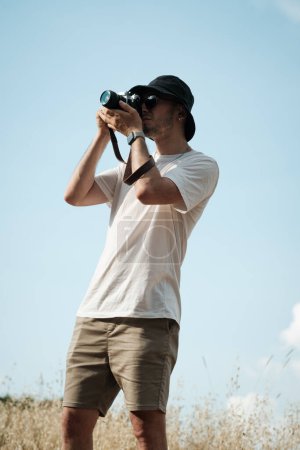 Photo for A portrait of a man in his twenties, casually dressed in a white t-shirt, beige shorts, sunglasses, and a black sailor cap, with a classic camera slung over his shoulder. Green hills and dry grass form the backdrop - Royalty Free Image