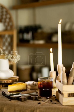 Foto de Junto a una rebanada de pastel de queso naranja, un Americano helado en un vaso se sienta en una tabla de madera sobre un mantel de arpillera, complementado por el cálido resplandor de las velas de fondo. - Imagen libre de derechos