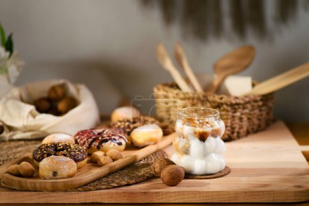 Photo for A wooden platter holds donuts, chocolate-filled cookies, and bite-sized cookies. Behind, walnuts rest on a white cloth, with wooden utensils in a wicker basket. An iced milk with espresso completes the scene - Royalty Free Image