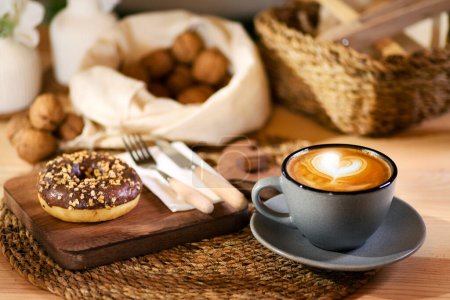 Foto de Un donut en una tabla de madera, al lado de un café con leche en una taza gris. Servilleta blanca con cubiertos de madera y nueces descascaradas en tela blanca en el fondo. - Imagen libre de derechos