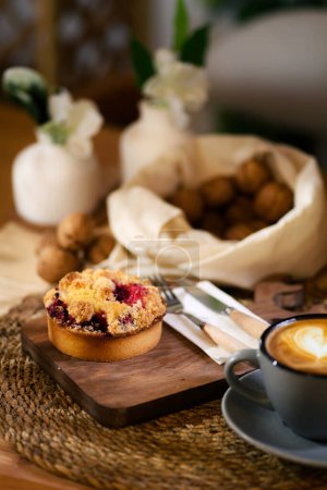Photo for On a wooden table, a gray cup of latte on a woven mat, beside a strawberry tart on a wood board, with knife and fork. Background of walnuts in cloth. - Royalty Free Image