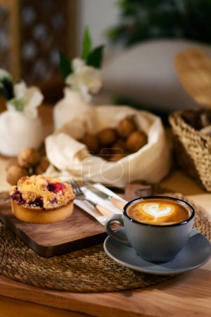 Téléchargez les photos : Sur une table en bois, une tasse grise de latte sur un tapis tissé, à côté d'une tarte aux fraises sur une planche de bois, avec couteau et fourchette. Fond de noix en tissu. - en image libre de droit
