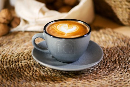 Foto de Un plano de cerca de un café con leche en una taza gris, sentado en una mesa de madera bañada en luz ambiental, irradiando calidez y comodidad. - Imagen libre de derechos