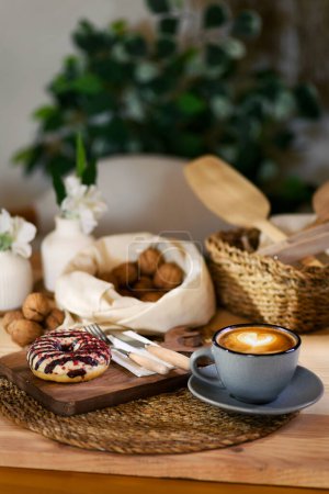 Photo for Front focus on a latte beside a donut presented on a wooden board. Background features walnuts in a white cloth and a wicker basket with wooden kitchen utensils, creating a warm, homely ambiance. - Royalty Free Image