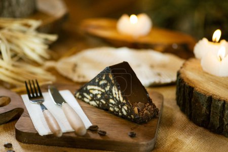 Photo for A slice of mosaic cake presented on burlap, alongside wooden cutlery and board. Captured with incense and warm candlelight. - Royalty Free Image