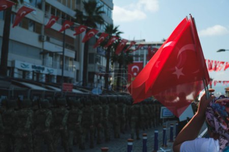 Foto de Esta llamativa fotografía tomada el 30 de agosto de 2023 en Izmir Kordon presenta prominentemente una bandera turca de colores vivos en primer plano. En el fondo, ligeramente fuera de foco debido a una profundidad de campo poco profunda, los soldados pueden ser vistos marchando en una c - Imagen libre de derechos