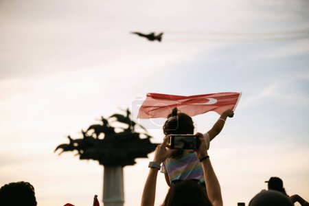 Téléchargez les photos : Izmir, Turquie, 9 septembre 2023,, place Cumhuriyet, Izmir, un enfant solitaire se distingue dans la foule, agitant le drapeau turc avec enthousiasme. La statue emblématique "Cumhuriyet Tree" est vue floue en arrière-plan, tandis que le ciel est orné de silh - en image libre de droit