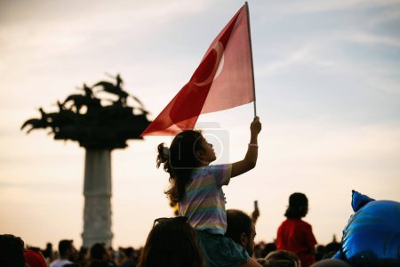 Foto de Izmir, Turquía, 9 de septiembre de 2023, en la Plaza Cumhuriyet, Izmir, un niño solitario se destaca entre la multitud, ondeando la bandera turca con entusiasmo. La icónica estatua de "Árbol Cumhuriyet" se ve borrosa en el fondo. - Imagen libre de derechos