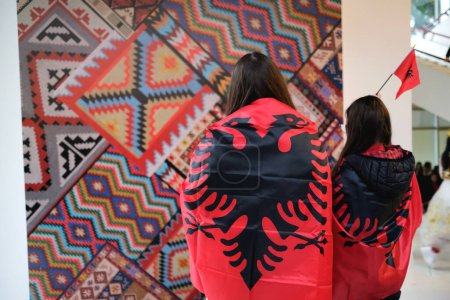 Photo for Tirana, Albania - November 28: Two women, their backs to the camera and wrapped in Albanian flags, pose in front of a traditional patterned backdrop - Royalty Free Image