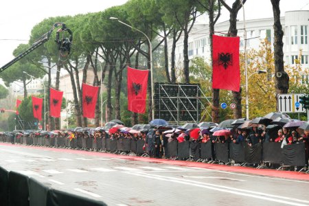 Foto de Tirana, Albania - 28 de noviembre de 2023: En un día lluvioso de la independencia, los ciudadanos con banderas y paraguas albaneses observan el desfile desde el borde de la carretera en la avenida de los mártires de la nación Boulevard avenida de los mártires de la nación - Imagen libre de derechos