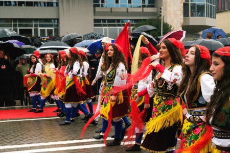 Téléchargez les photos : Tirana, Albanie - 28 novembre 2023 : Sur l'avenue des martyrs de la nation, près du palais Mère Teresa, les étudiants en tenue traditionnelle défilent avec enthousiasme le jour de l'indépendance pluvieuse, avec des spectateurs portant des parapluies et des drapeaux albanais dans le backgrou - en image libre de droit