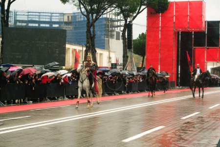 Foto de Tirana, Albania - 28 de noviembre de 2023: Frente al Palacio de la Madre Teresa en el Boulevard de los Mártires de la Nación, el representante Skanderbeg monta un caballo blanco y es observado por los ciudadanos durante un lluvioso Día de la Independencia.. - Imagen libre de derechos