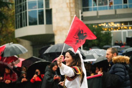 Foto de Tirana, Albania - 28 de noviembre de 2023: En la Avenida de los Mártires de la Nación, cerca del Palacio de la Madre Teresa, una mujer vestida de manera tradicional camina jubilosamente con una bandera albanesa en un Día de la Independencia lluviosa, con espectadores sosteniendo sombrillas y banderas en el bac - Imagen libre de derechos