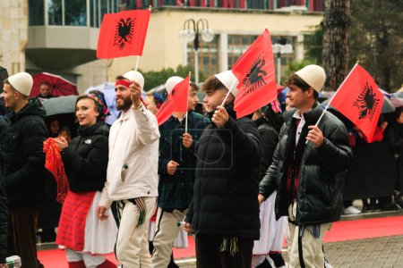 Photo for Tirana, Albania - November 28, 2023: On Avenue of Martyrs of the Nation, near Mother Teresa Palace, students in traditional attire march with Albanian flags in hand on a rainy Independence Day, while onlookers with umbrellas and flags watch in the ba - Royalty Free Image