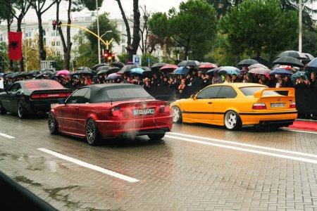 Téléchargez les photos : Tirana, Albanie - 28 novembre 2023 : Sur l'avenue des martyrs de la nation, les citoyens regardent un défilé automobile modifié le jour de l'indépendance pluvieux, avec une BMW 3 E46 rouge et jaune et un challenger de l'esquive de couleur noire étiqueté Cair Tuning Team - en image libre de droit