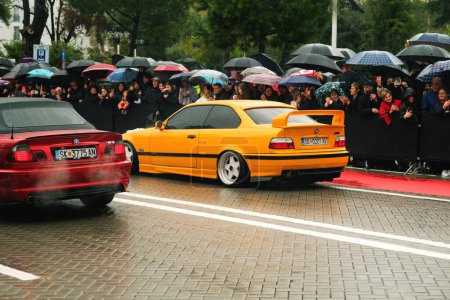 Photo for Tirana, Albania - November 28, 2023: On Avenue of Martyrs of the Nation, citizens watch a modified car parade on a rainy Independence Day, featuring a red and yellow BMW 3 E46 and labeled Cair Tuning Team - Royalty Free Image