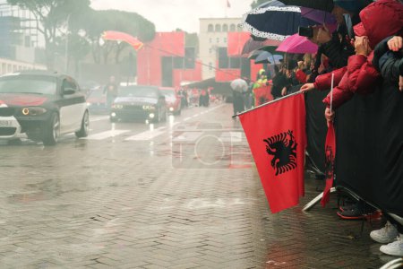 Téléchargez les photos : Tirana, Albanie - 28 novembre 2023 : Sur l'avenue des martyrs de la nation, les citoyens regardent un défilé de voitures modifiées le jour de l'indépendance pluvieux, avec une BMW rouge au premier plan et un drapeau albanais. - en image libre de droit