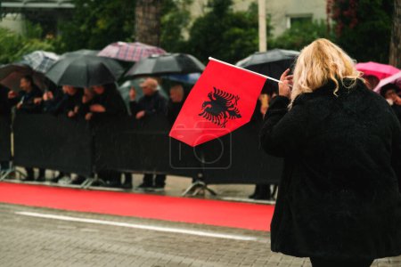 Foto de Tirana, Albania - 28 de noviembre de 2023: Frente al Palacio de la Madre Teresa en la Avenida de los Mártires de la Nación, una mujer camina jubilosamente con una bandera albanesa en un Día de la Independencia lluviosa, con espectadores sosteniendo paraguas y banderas en el fondo - Imagen libre de derechos