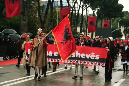 Téléchargez les photos : Tirana, Albanie - 28 novembre 2023 : Sur l'avenue des martyrs de la nation, étudiants et personnes en tenue traditionnelle défilent avec enthousiasme avec des drapeaux albanais le jour de l'indépendance pluvieux, avec des spectateurs tenant des parapluies et des drapeaux en arrière-plan. - en image libre de droit
