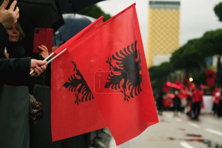 Foto de Tirana, Albania - 28 de noviembre de 2023: En la Avenida de los Mártires de la Nación, los espectadores observan con entusiasmo las actuaciones del Día de la Independencia bajo la lluvia, sosteniendo banderas albanesas, con un primer plano de la bandera en la foto. - Imagen libre de derechos