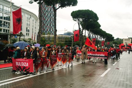 Photo for Tirana, Albania - November 28, 2023: On Avenue of Martyrs of the Nation, students and people in traditional attire enthusiastically march with Albanian flags on a rainy Independence Day, with spectators holding umbrellas and flags in the background. - Royalty Free Image