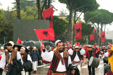 Foto de Tirana, Albania - 28 de noviembre de 2023: En la avenida de los Mártires de la Nación, estudiantes y personas vestidas con ropa tradicional marchan con entusiasmo con banderas albanesas en un día de la independencia lluviosa, con los espectadores sosteniendo sombrillas y banderas en el fondo. - Imagen libre de derechos
