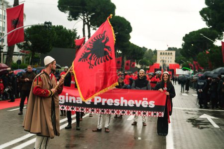 Foto de Tirana, Albania - 28 de noviembre de 2023: En la avenida de los Mártires de la Nación, estudiantes y personas vestidas con ropa tradicional marchan con entusiasmo con banderas albanesas en un día de la independencia lluviosa, con los espectadores sosteniendo sombrillas y banderas en el fondo. - Imagen libre de derechos