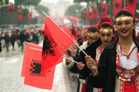 Foto de Tirana, Albania - 28 de noviembre de 2023: En la Avenida de los Mártires de la Nación, durante un Día de la Independencia lluviosa, las estudiantes marchan en fila, sonriendo y sosteniendo banderas albanesas con entusiasmo. - Imagen libre de derechos